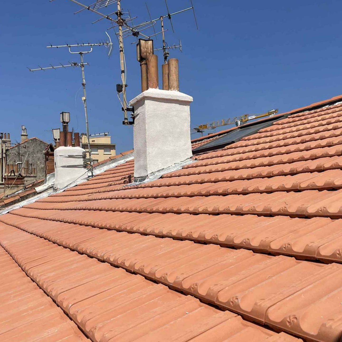 Toit de tuiles rouges, cheminée blanche et antennes se détachant sur un ciel bleu limpide.