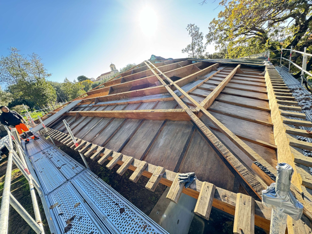 Toiture en construction ; poutres et planches de bois, échafaudages et un ouvrier en uniforme orange sur fond de ciel ensoleillé.
