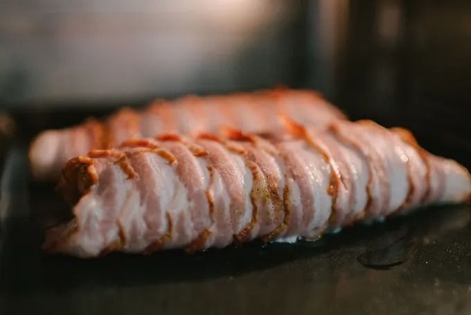 Mit Speck umwickelte Lebensmittel auf einem dunklen Backblech im Ofen, teilweise sichtbar.