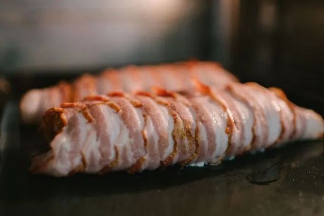 Mit Speck umwickelte Lebensmittel auf einem dunklen Backblech im Ofen, teilweise sichtbar.