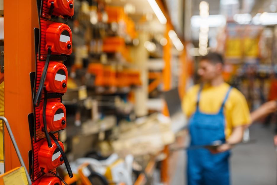 Cintas métricas expuestas en un estante en una ferretería, con un empleado con mono azul al fondo.