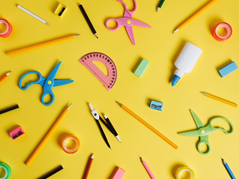 Fondo amarillo con material escolar disperso: lápices, tijeras, pegamento, cinta adhesiva, transportador.