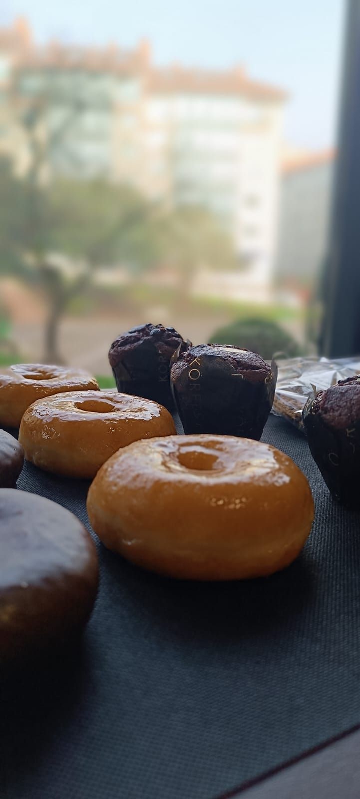 Un montón de donas y brownies están sobre una mesa frente a una ventana.