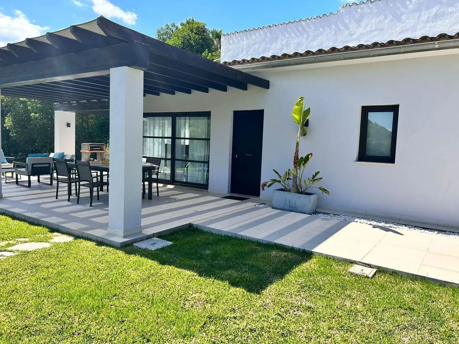 Patio de una casa moderna de color blanco con zona de estar cubierta, puertas de cristal y un césped verde bañado por la luz del sol.