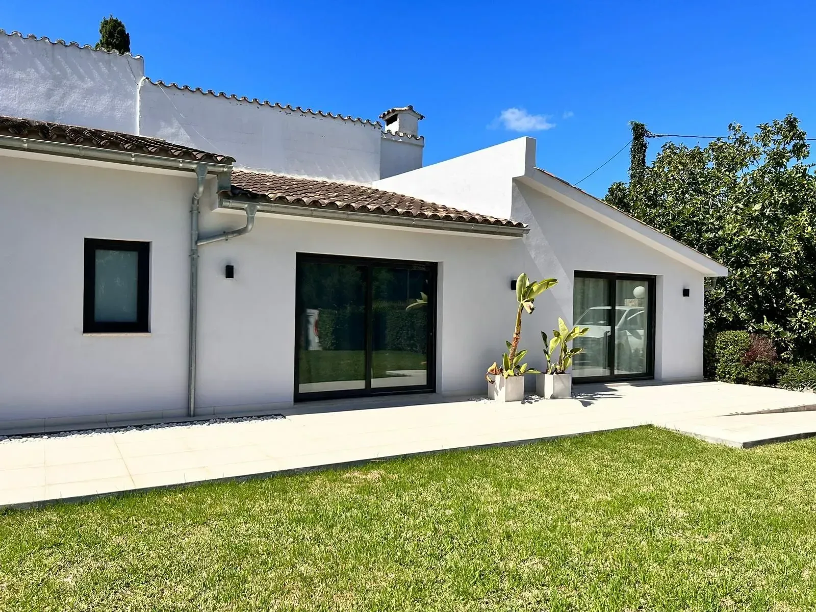 Exterior de una casa moderna blanca con ventanas de marcos negros, patio y césped verde bajo un cielo azul.