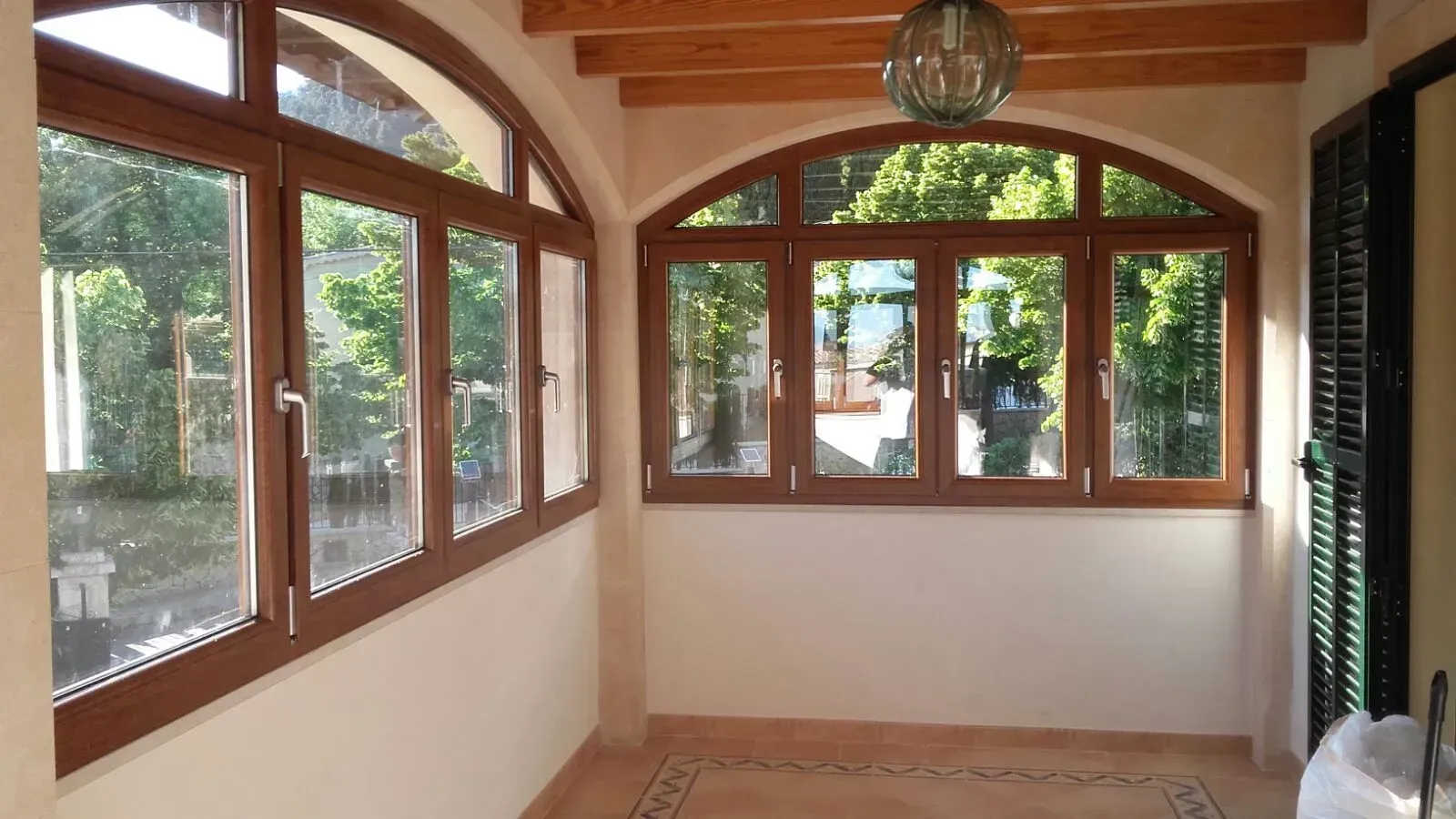 Terraza acristalada vacía con techo de vigas de madera, grandes ventanales arqueados y paredes blancas.