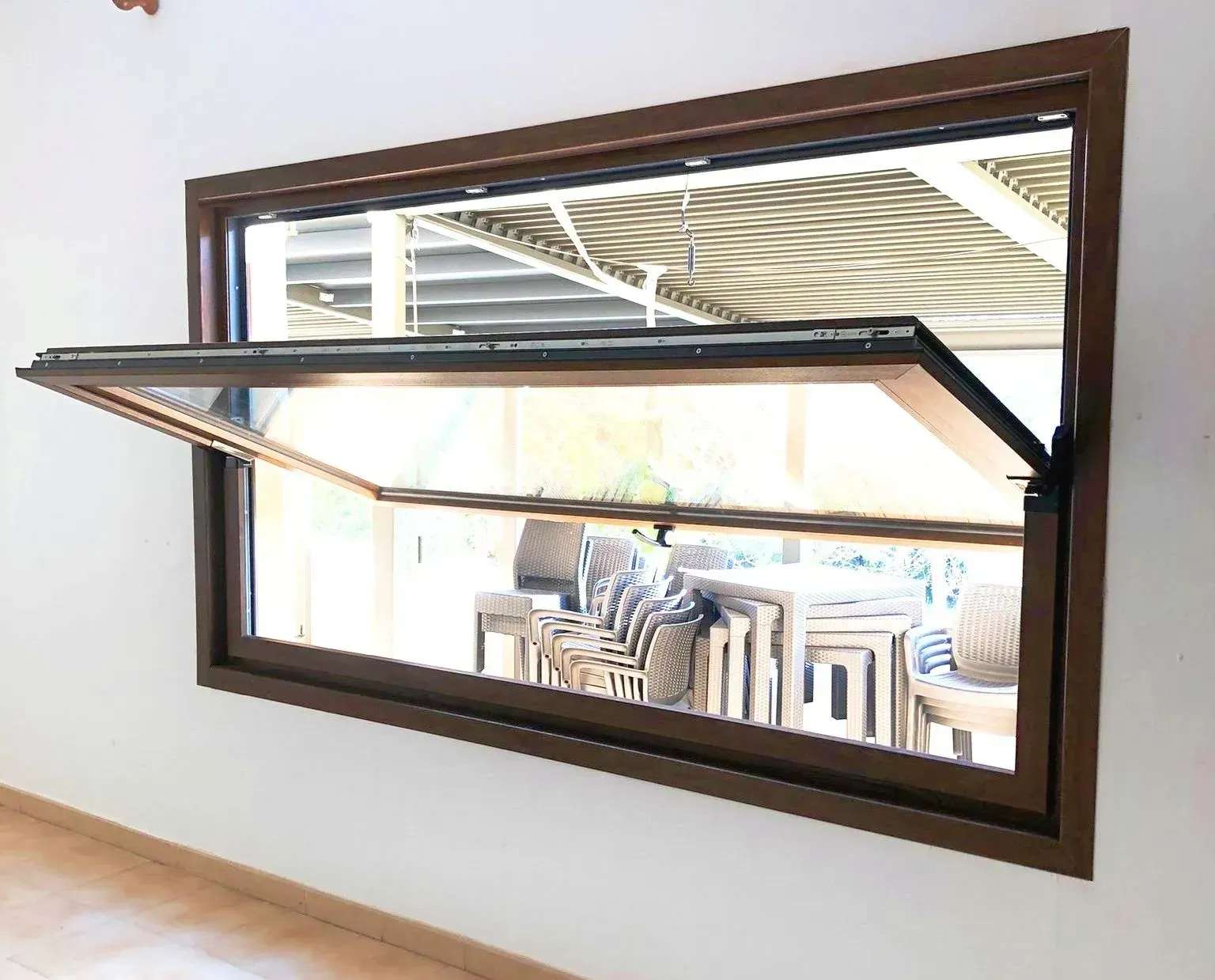 Ventana abierta que da a un patio con sillas a rayas, enmarcado por molduras marrones y un toldo abatible.