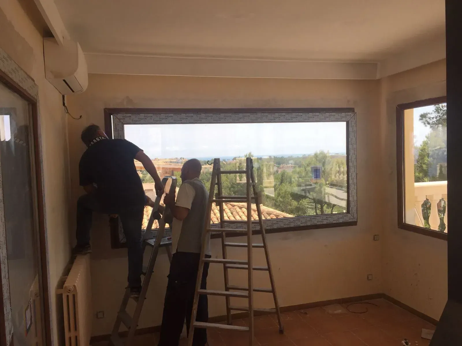 Dos trabajadores subidos a escaleras instalan una gran ventana en una habitación luminosa.