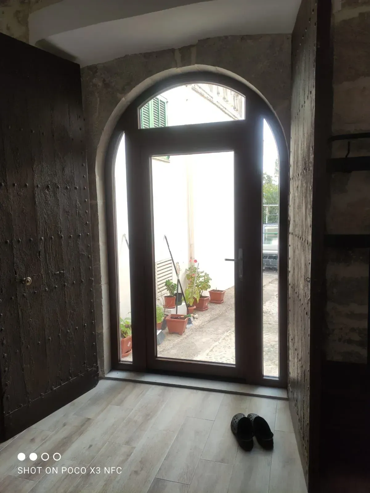 Entrada con poca luz, puerta de cristal arqueada, plantas en macetas en el exterior y zapatos negros en el suelo.