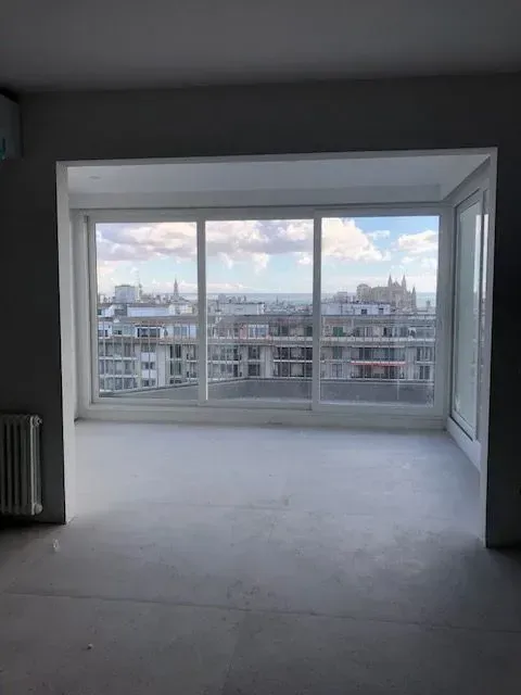 Habitación vacía sin amueblar con un gran ventanal con vistas a los edificios de la ciudad y al cielo nublado.
