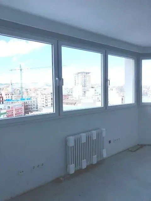 Habitación vacía con grandes ventanales que dan al horizonte de la ciudad y un radiador blanco debajo.