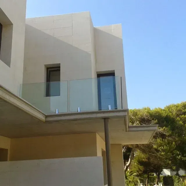 Casa moderna de color beige con balcón de cristal, bajo un cielo azul claro, junto a árboles.