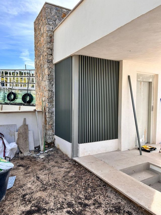 Patio parcialmente terminado con muro de piedra, listones verticales y herramientas de construcción junto a una casa moderna blanca.