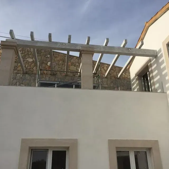 Balcón con techo de piedra sobre dos ventanas en un edificio de color crema, con vigas de pérgola de madera en la parte superior.