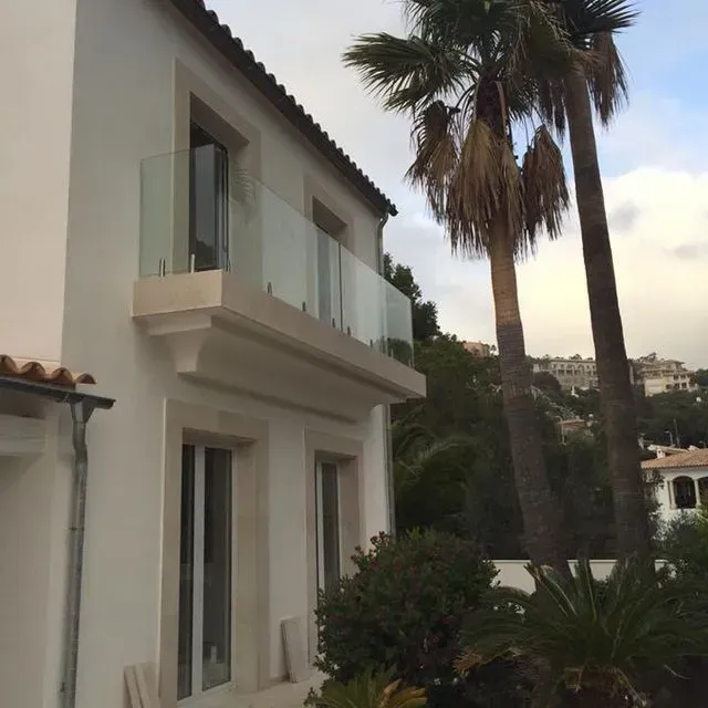 Casa moderna blanca con balcón, altas palmeras y jardín en una ladera costera.