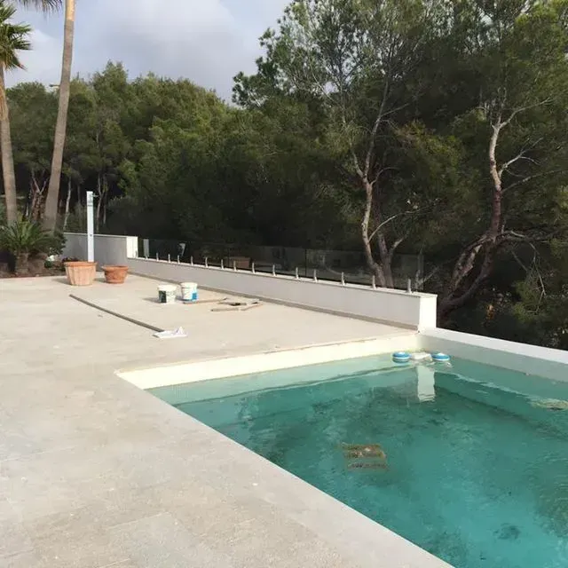 Piscina exterior con agua turquesa junto a una terraza de hormigón y árboles detrás de un muro bajo.