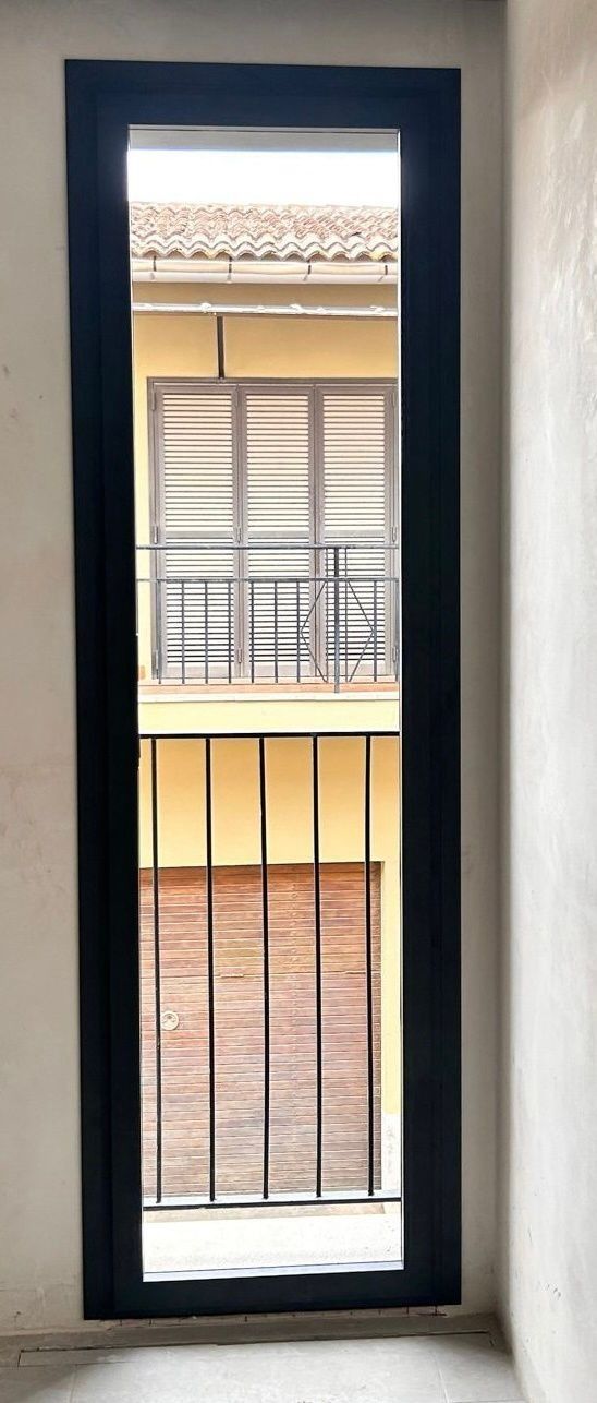 Ventana alta y estrecha con marco negro y barrotes de metal que da a la fachada de un edificio iluminado por el sol.