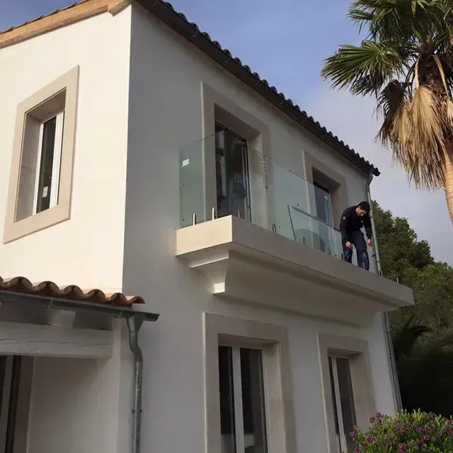 Casa blanca de dos plantas con balcón de cristal, y una persona de pie en el balcón superior cerca de una palmera.