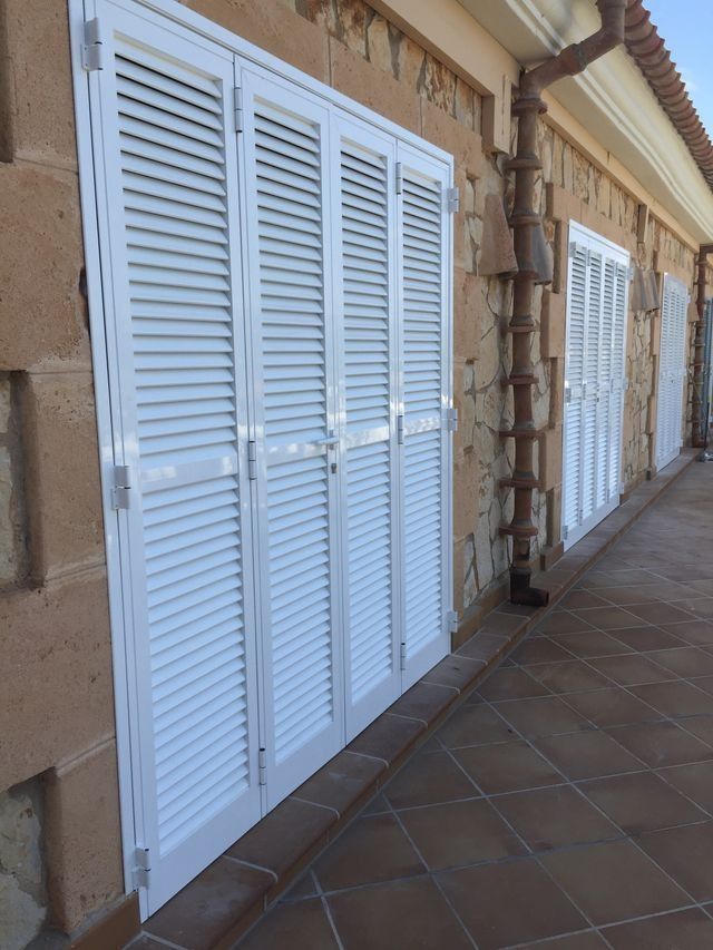Puertas blancas con contraventanas a lo largo de un porche soleado con baldosas, junto a una pared de estuco.