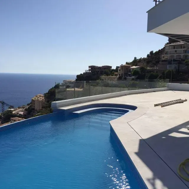 Piscina infinita con vistas al océano y villas en la ladera bajo un cielo azul despejado.