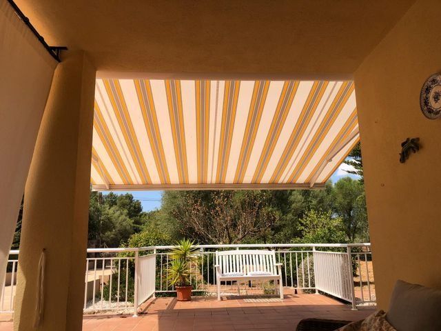 Vista del patio con toldo a rayas, barandilla blanca, plantas en macetas y vegetación al fondo.