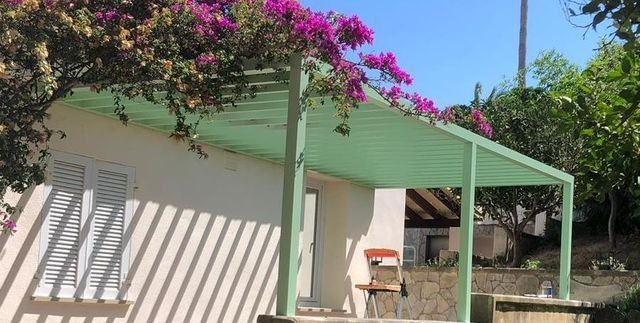 Casa blanca con toldo y porche verdes, a la sombra de buganvillas rosas en un jardín soleado.