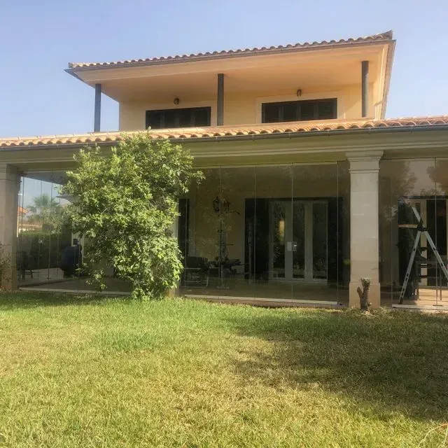 Casa de dos plantas con patio cubierto, césped y una escalera apoyada cerca de la entrada.