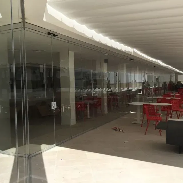Terraza de cafetería con mesas y sillas rojas bajo una pasarela cubierta junto a paredes de cristal.