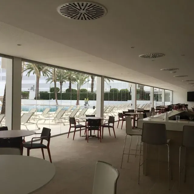 Cafetería al aire libre junto a la piscina con mesas, sillas y palmeras más allá de los grandes ventanales.