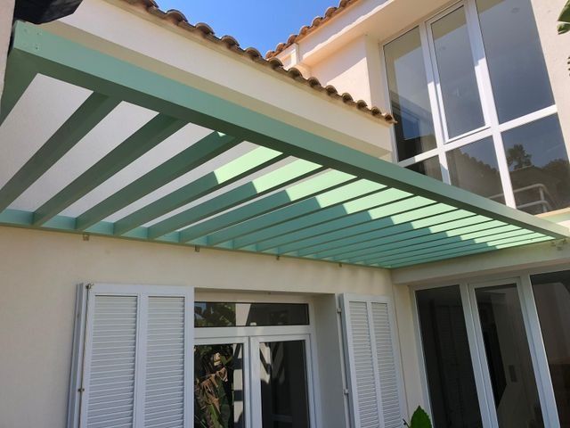 Patio con toldo de rayas verdes que se extiende desde una casa blanca con puertas y ventanas con contraventanas.
