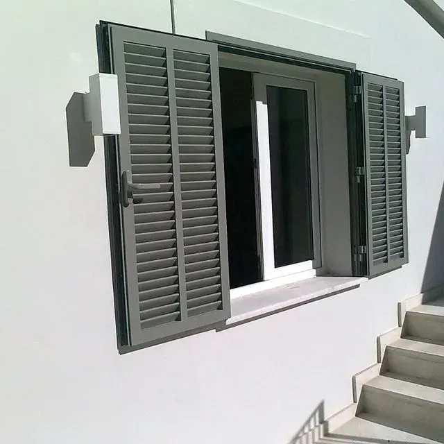 Persianas grises abiertas en una pared blanca, dejando ver una ventana abierta junto a las escaleras exteriores.