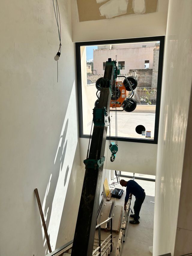 Un obrero de la construcción se encuentra en una escalera utilizando maquinaria cerca de una ventana, con otro trabajador debajo.