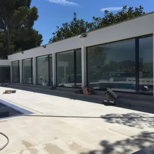 Moderna casa blanca con puertas de cristal del suelo al techo junto a una terraza con piscina bañada por el sol.