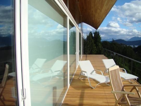 Terraza moderna de madera con tumbonas blancas, puertas de cristal y vistas a la montaña bajo un cielo azul.