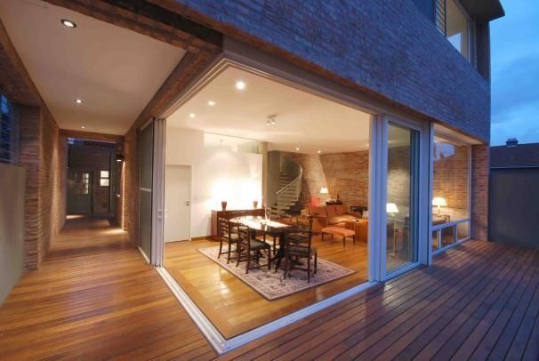 Patio de casa moderna con puertas corredizas de vidrio abiertas, área de comedor iluminada y terraza de madera al atardecer.