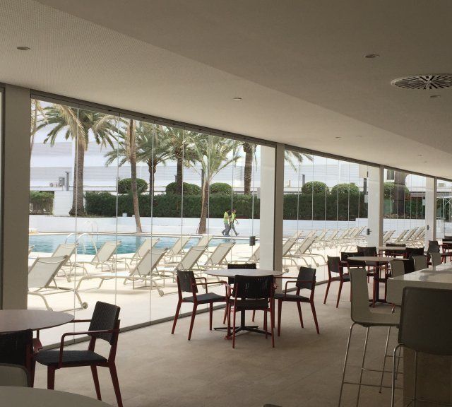 Salón interior con vistas a una piscina con tumbonas blancas y palmeras en el exterior.