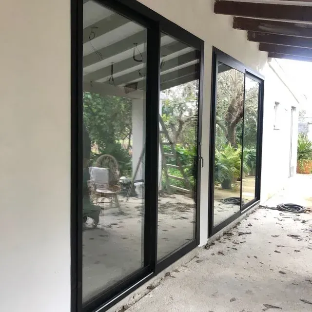 Pasarela cubierta junto a puertas de cristal con marcos negros que dan a un patio frondoso con sillas de exterior.