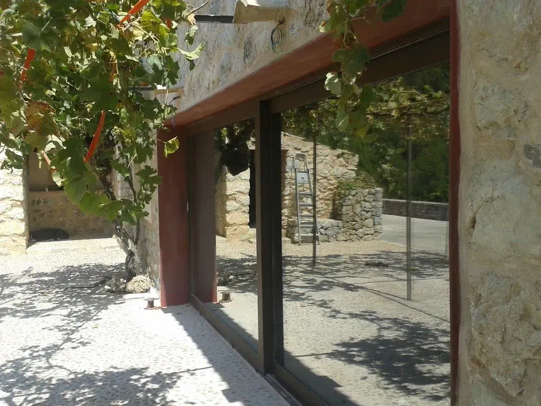 Escaparate de cristal con detalles rojos junto a una pared de estuco y sombras de árboles frondosos en la acera.