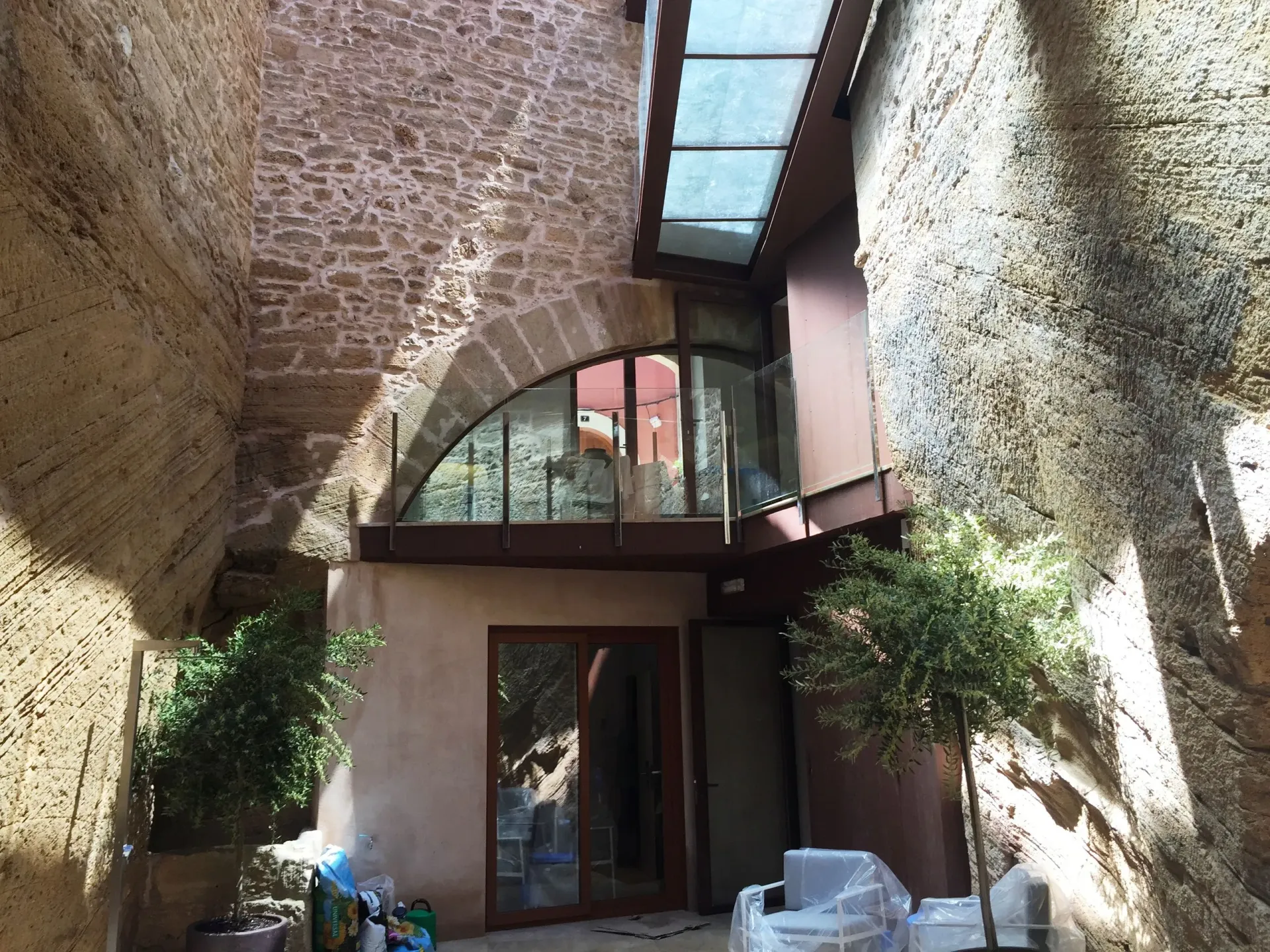Patio estrecho de piedra con abertura arqueada, techo de cristal y plantas en macetas alrededor de una puerta.