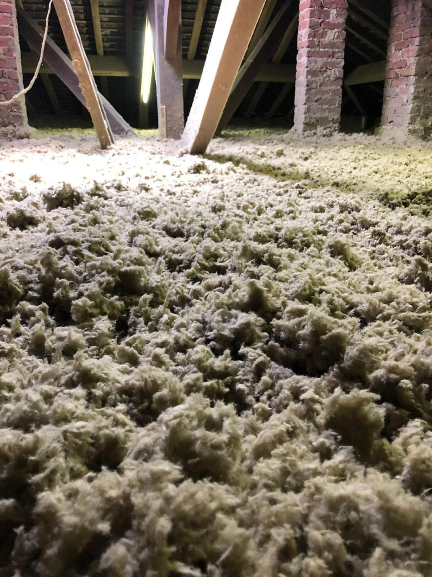 Grenier avec isolation lâche et fibreuse, poutres apparentes et colonnes en briques, éclairé par un luminaire.