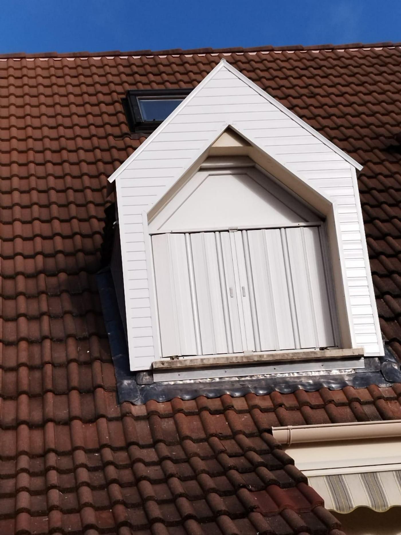 Lucarne blanche sur un toit de tuiles brunes sous un ciel bleu.