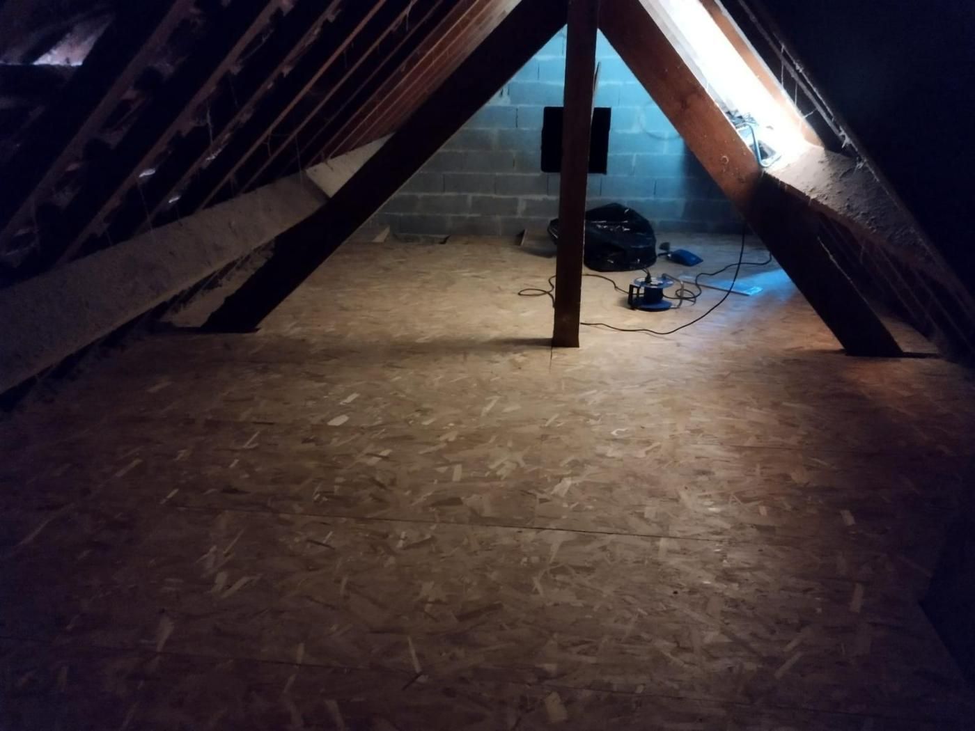 Grenier avec plancher en contreplaqué, poutres en bois et mur en béton. Câbles et équipements électriques visibles.