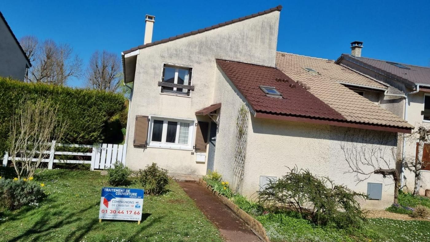 Maison avec extérieur blanc cassé, toit et volets marron, un panneau de vente sur la pelouse et un ciel bleu.