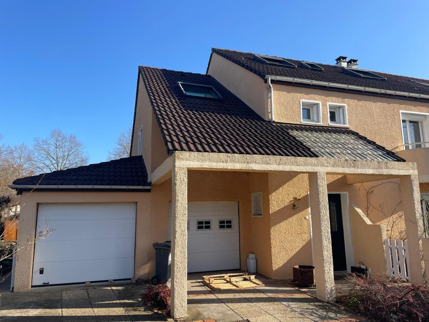 Maison à deux étages avec extérieur en stuc beige, toit foncé, garage et porche ouvert par une journée ensoleillée.