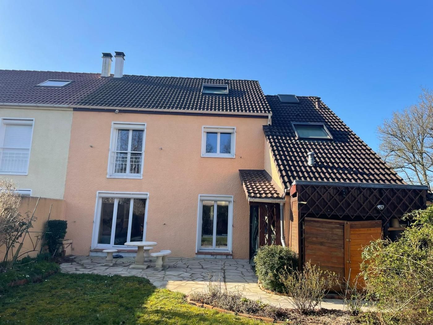 Arrière d'une maison à deux étages avec un extérieur couleur pêche, un patio en pierre et une pelouse verte sous un ciel bleu.
