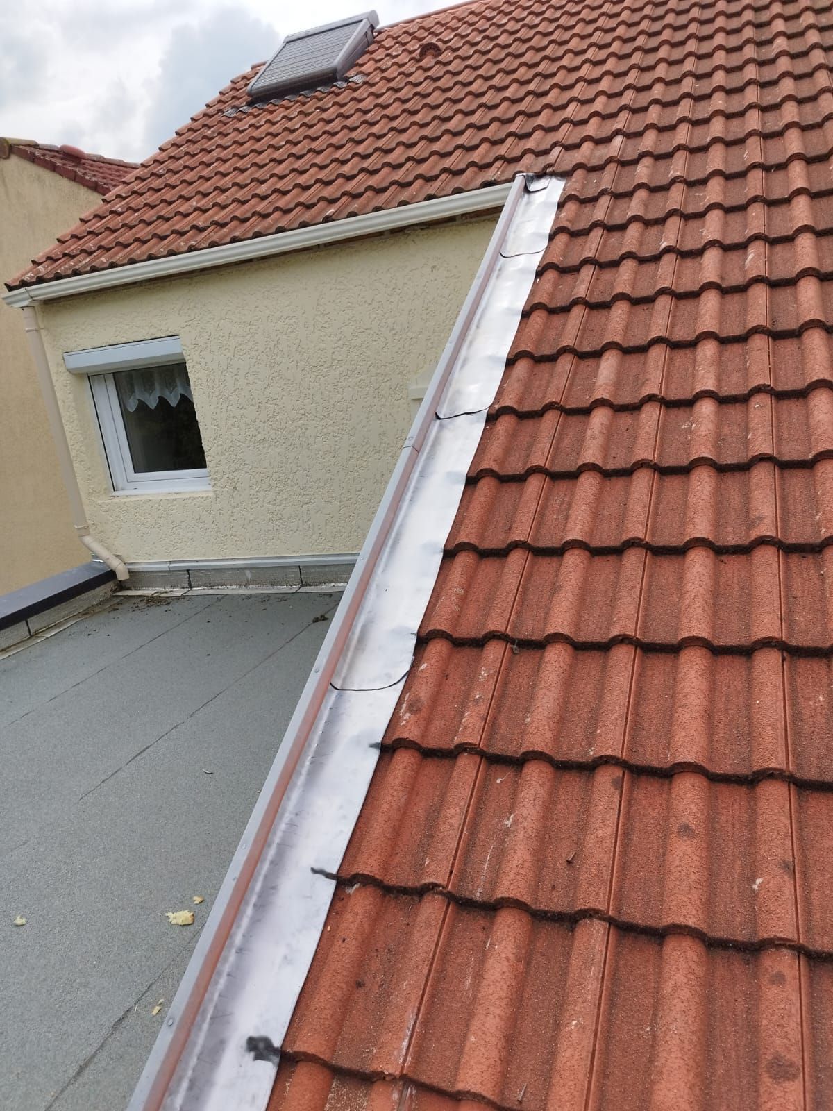 Toit en tuiles rouges avec gouttière métallique, jouxtant un toit plat et un mur en stuc avec une fenêtre