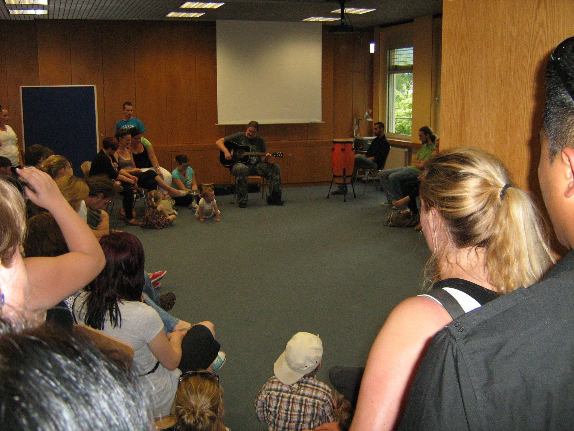 Eine Gruppe von Menschen, die in einem Raum sitzen und einem Mann beim Gitarrenspielen zuschauen