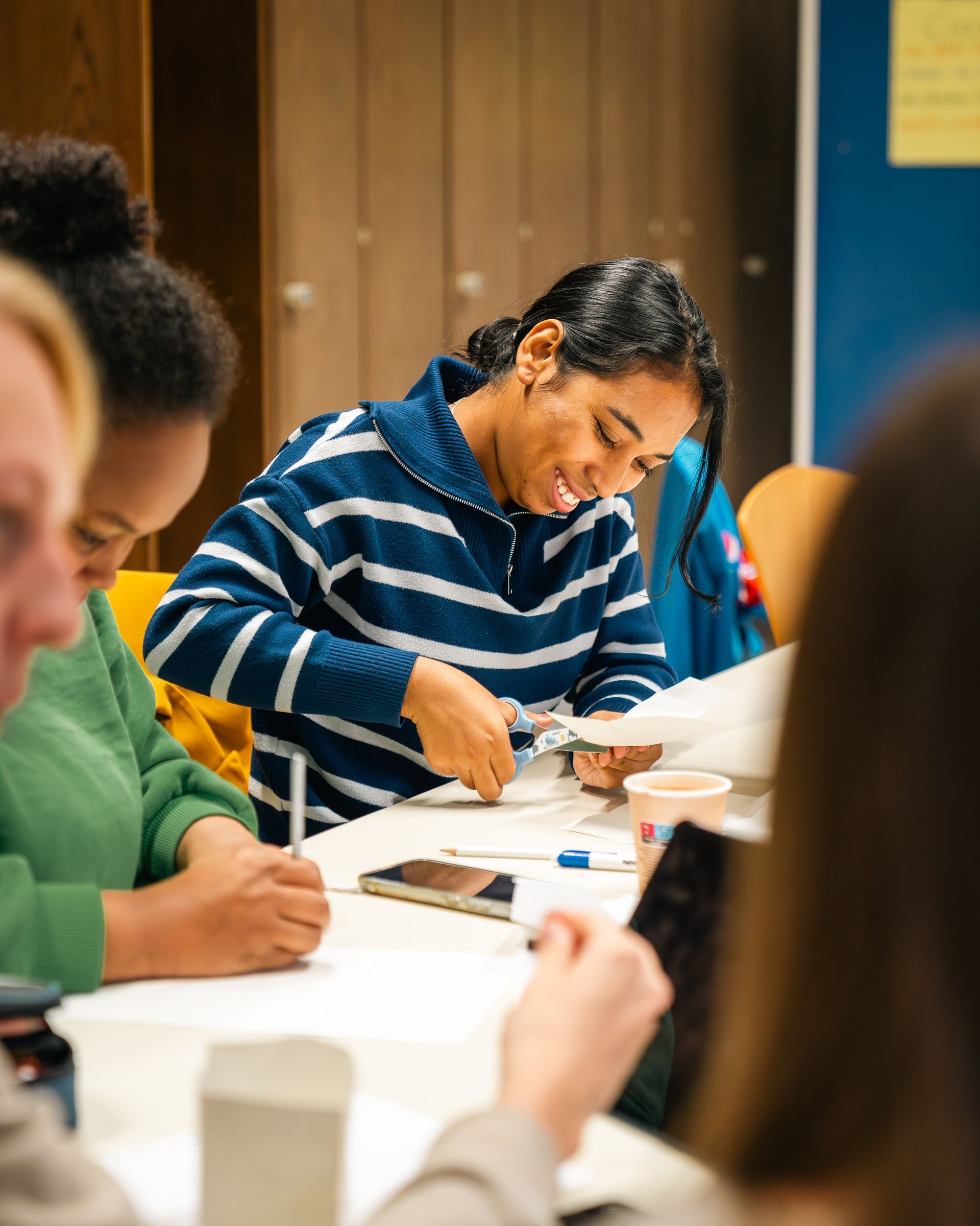 Menschen arbeiten gemeinsam an einem Tisch. Eine Frau im blau-weiß gestreiften Hemd schneidet lächelnd Papier. Andere schreiben und arbeiten mit Geräten.