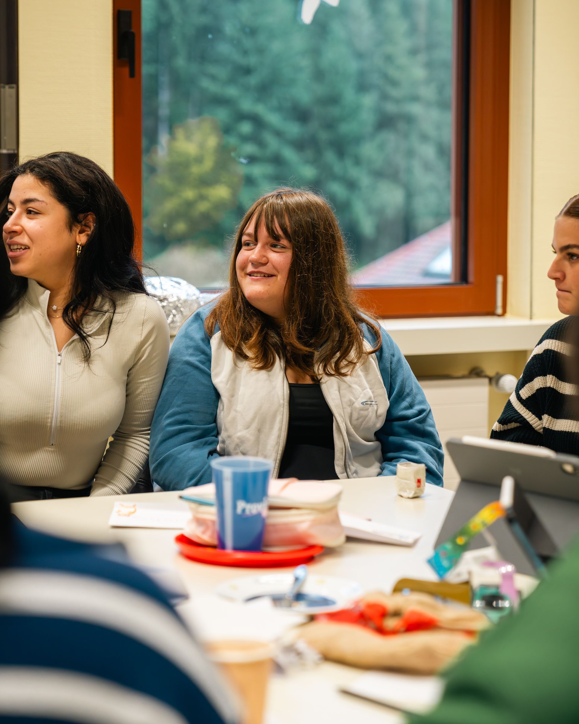 Menschen sitzen an einem Tisch. Eine Frau in blauer Jacke lächelt. Sonnenlicht fällt im Hintergrund durch ein Fenster.