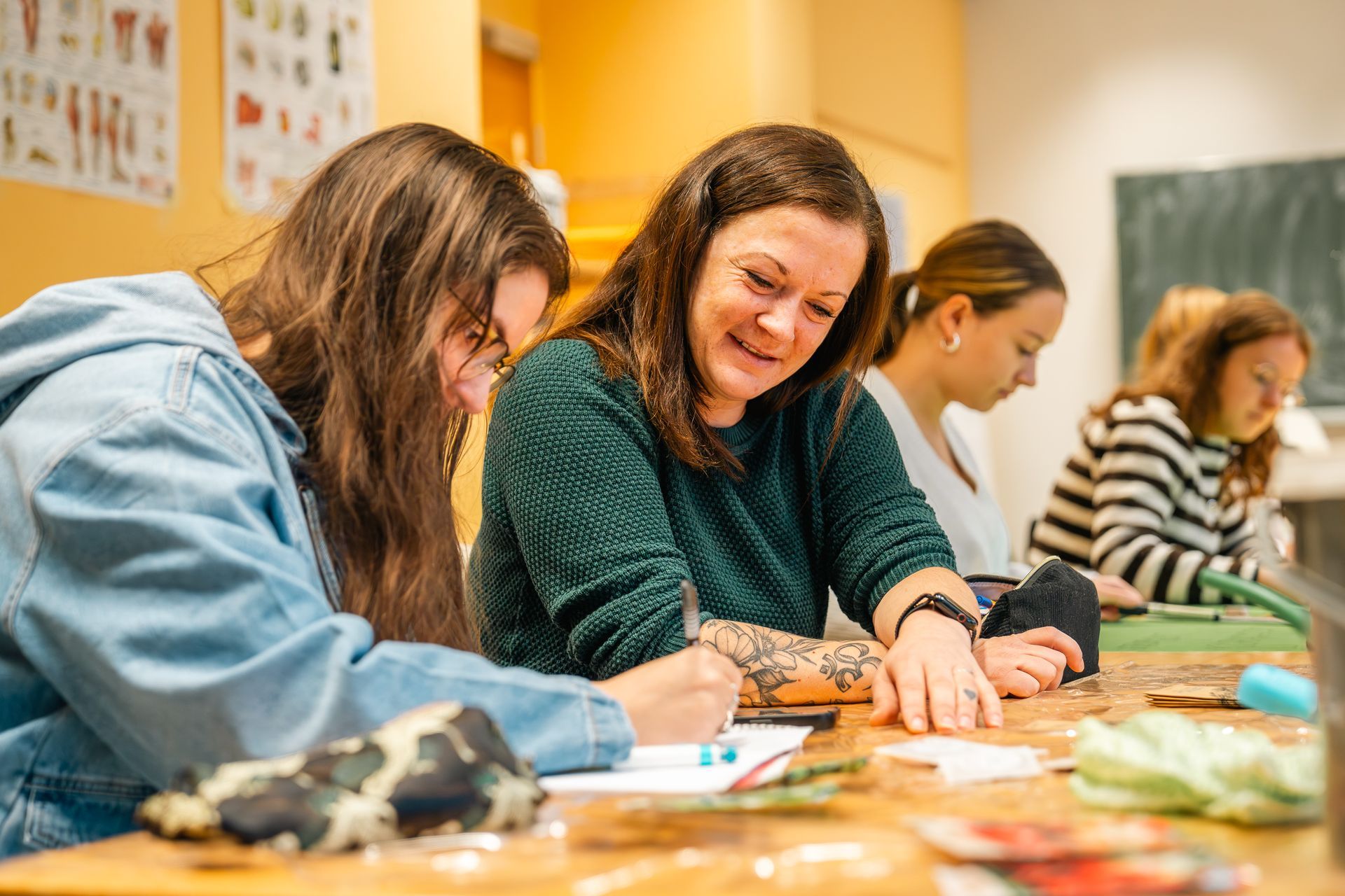 Vier Personen sitzen an einem Tisch und basteln. Eine Frau im grünen Pullover lächelt, während sie auf ein Blatt Papier schaut.